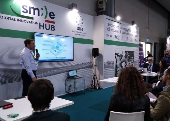 Foto di uno stand fieristico con presentazione su schermo e pubblico seduto in ascolto.