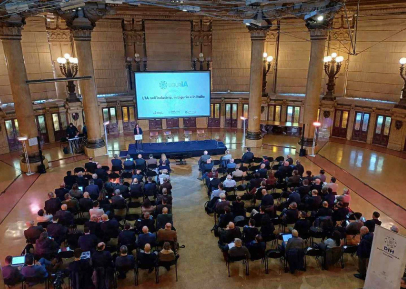 Sala conferenze con pubblico seduto e palco con schermo.