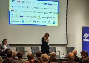 Relatrice sul palco durante l'evento SPS Italia On Tour a Genova sull’innovazione industriale, con pubblico in sala e slide proiettata sullo schermo.